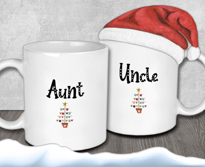Two white mugs with the text &quot;Aunt&quot; and &quot;Uncle&quot; printed on them, along with a small Christmas tree design. A Santa hat is placed on top of one of the mugs.