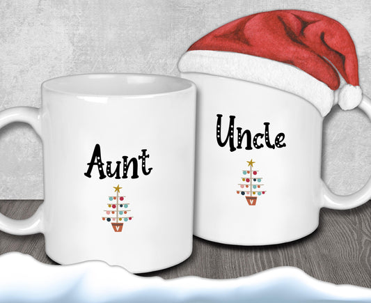 Two white mugs with the text &quot;Aunt&quot; and &quot;Uncle&quot; printed on them, along with a small Christmas tree design. A Santa hat is placed on top of one of the mugs.