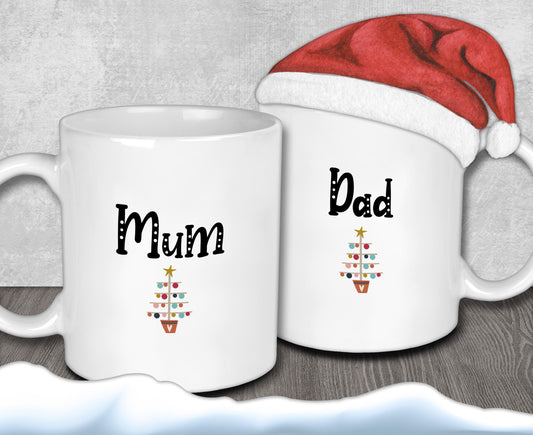 Two white mugs with Christmas tree designs and the names "Mum" and "Dad" printed on them, along with a Santa hat.