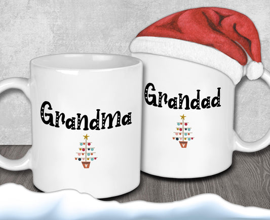 Two white mugs with the text &quot;Grandma&quot; and &quot;Granddad&quot; printed on them, along with a small Christmas tree design. A Santa hat is placed on top of one of the mugs.