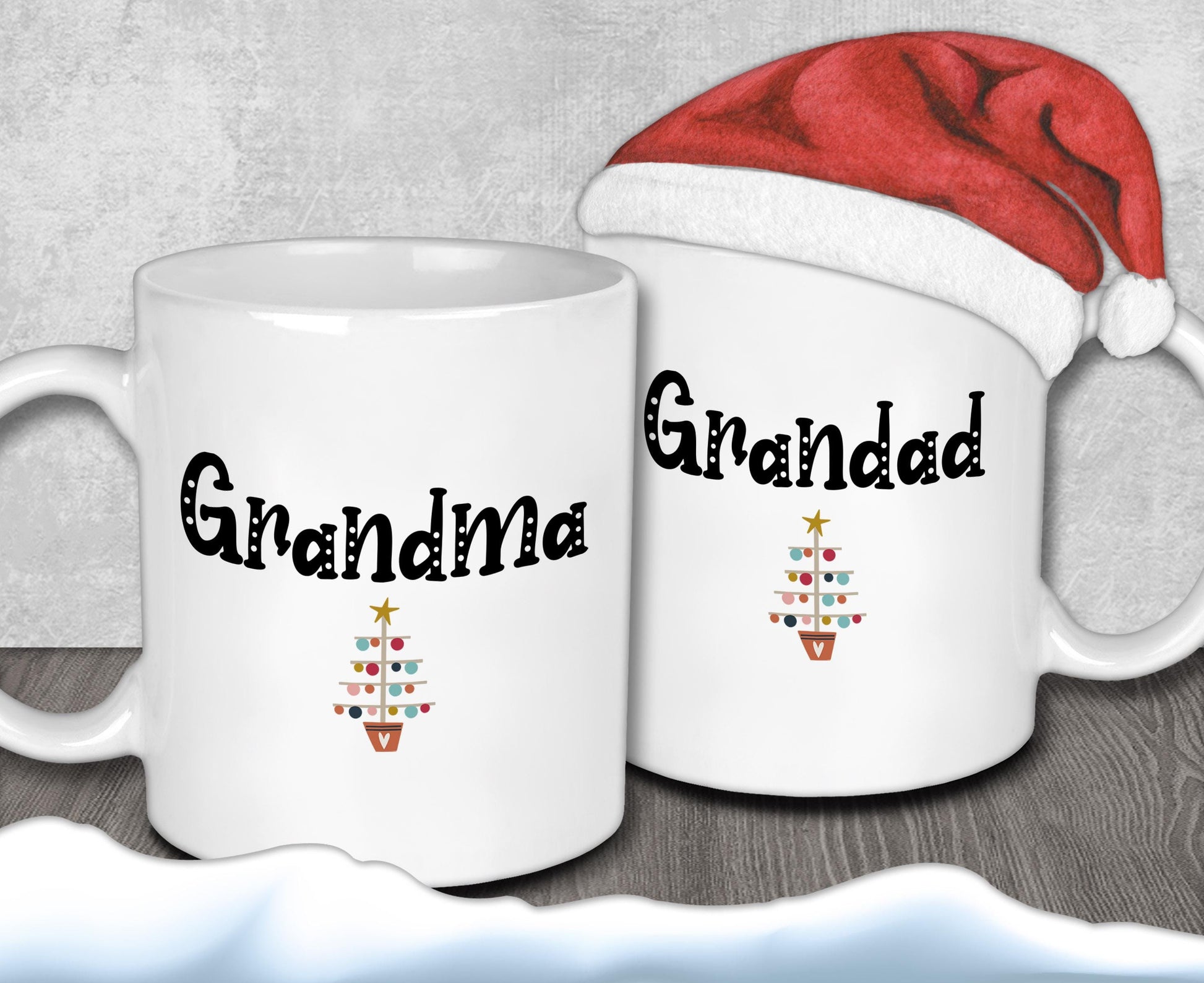 Two white mugs with the text "Grandma" and "Granddad" printed on them, along with a small Christmas tree design. A Santa hat is placed on top of one of the mugs.