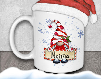 A white mug with a red and white striped gnome design and the name "Nonna" printed on it, along with snowflakes and a Santa hat.