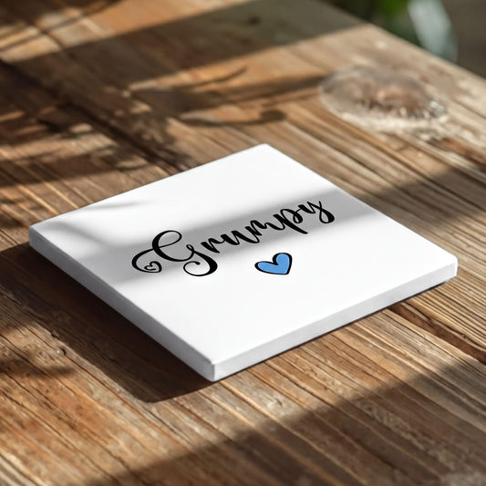 A white square coaster with the word "Grumpy" written on it, along with a blue heart symbol, placed on a wooden surface.