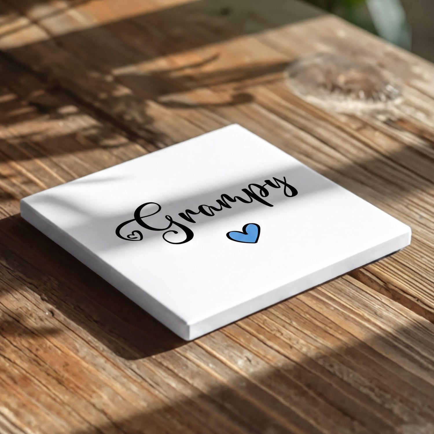 A white square coaster with the word "Grumpy" written on it, along with a blue heart symbol, placed on a wooden surface.