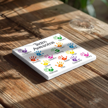 A white square coaster with colorful handprint designs and the text "Team Awesome" printed on it, placed on a wooden surface.