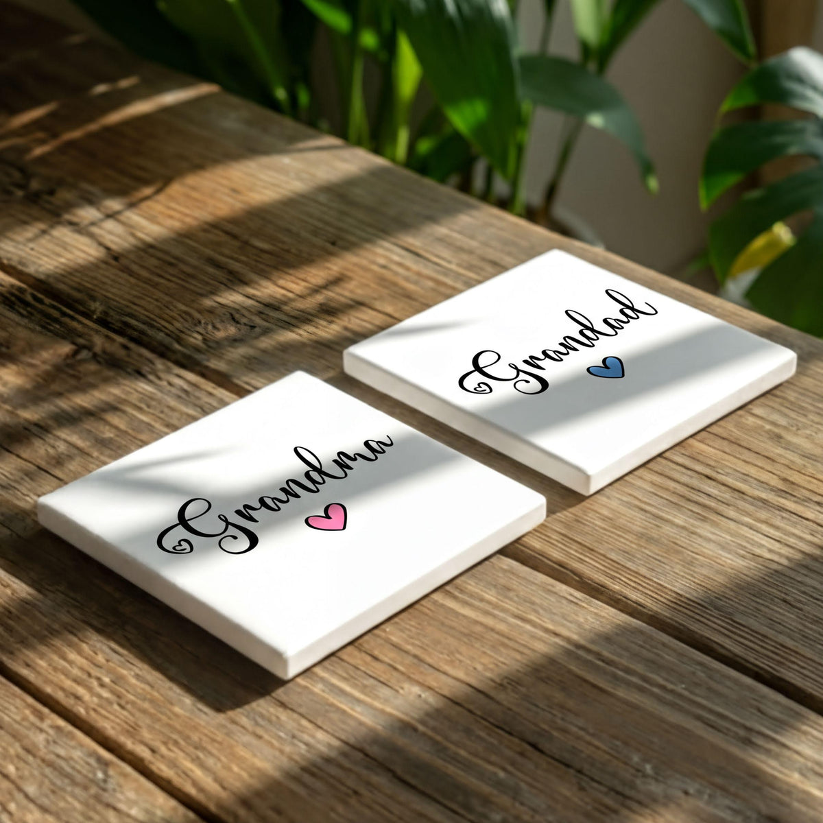 Two white square coasters with the word "Grandma" written on them, along with a heart symbol, placed on a wooden surface.