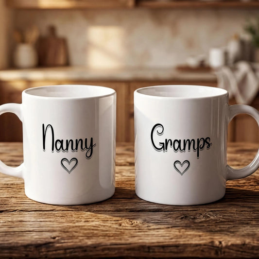 Two white mugs with black text that says "Nanny" and "Grams" on them, placed on a wooden table in a kitchen setting.