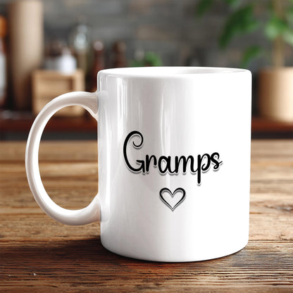 A white ceramic mug with the text "Gramps" and a heart symbol printed on it, placed on a wooden surface.