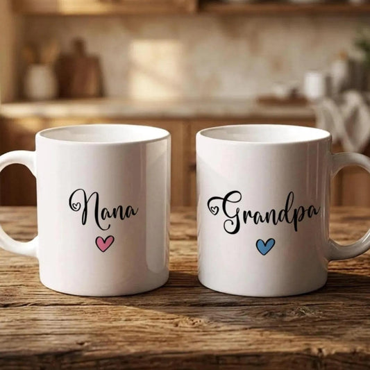 Two white mugs with the names "Nana" and "Grandpa" printed on them, along with hearts, are placed on a wooden surface.