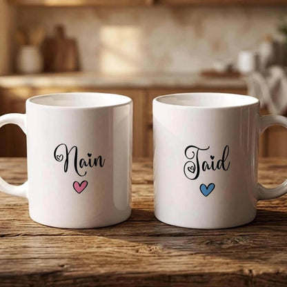 Two white mugs with the names "Nain" and "Taid" printed on them, along with a heart symbol, are placed on a wooden surface.