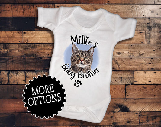 A white baby onesie with a cat image and the text "Milly's Baby Brother" printed on it, placed on a wooden surface.