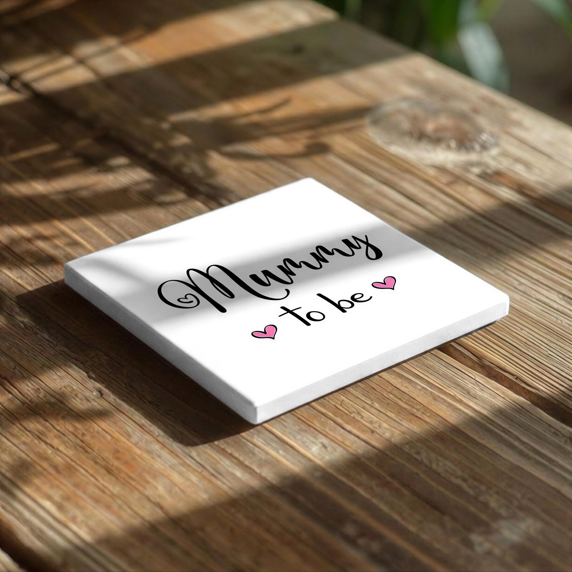 A white square with the text "Mummy to be" written on it, along with two pink heart symbols, resting on a wooden surface.