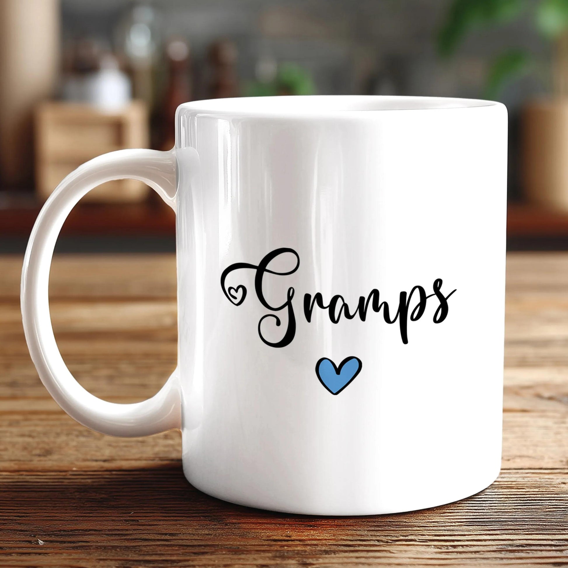 A white ceramic mug with the text "Gramps" and a blue heart symbol printed on it, placed on a wooden surface.