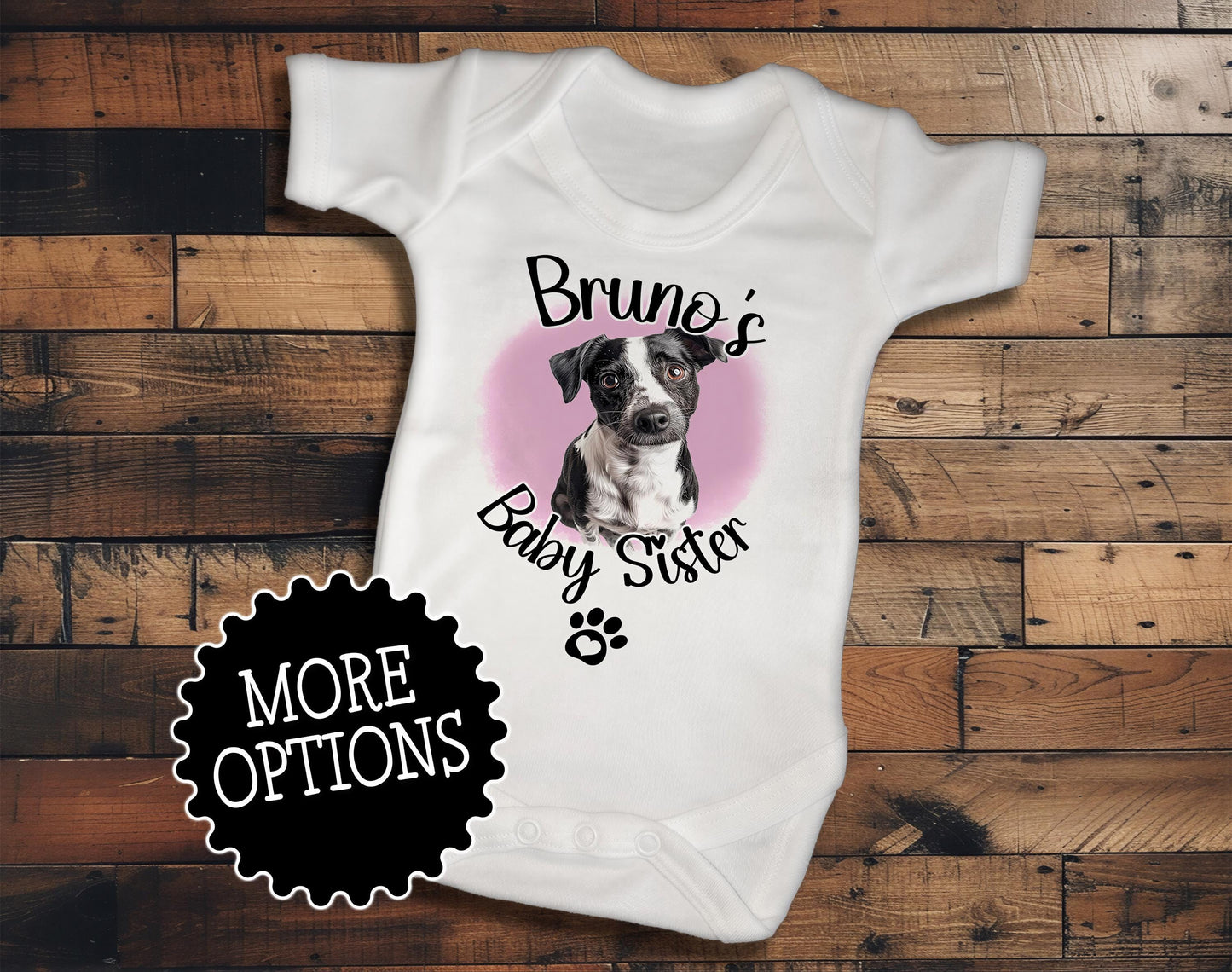 A white baby onesie with a graphic of a dog and the text "Bruno's Baby Sister" printed on it, placed on a wooden surface.