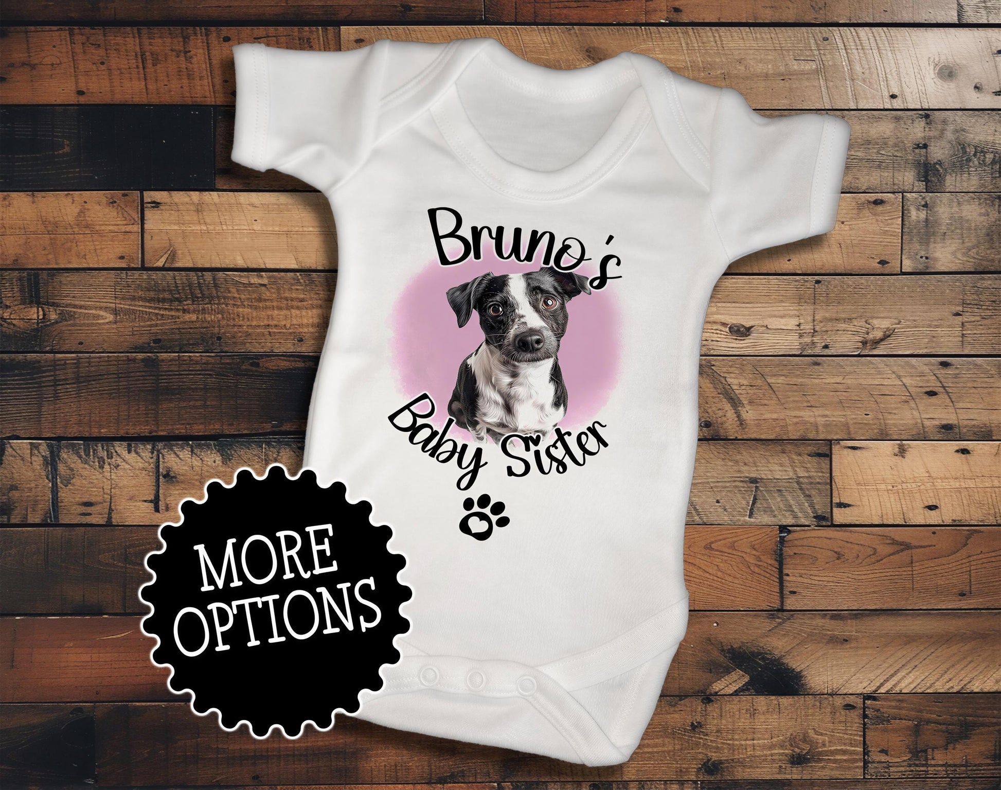 A white baby onesie with a graphic of a dog and the text "Bruno's Baby Sister" printed on it, placed on a wooden surface.