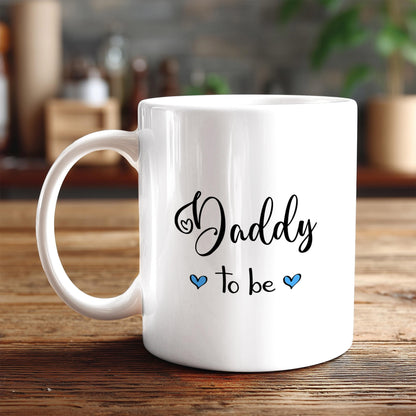 A white ceramic mug with the text "Daddy to be" printed on it, along with two blue heart symbols.