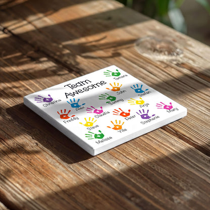 A white square coaster with colorful handprint designs and the text "Team Awesome" printed on it, placed on a wooden surface.