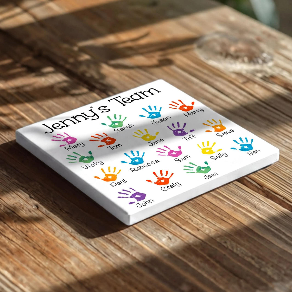 A white square coaster with colorful handprint designs on it, placed on a wooden surface.