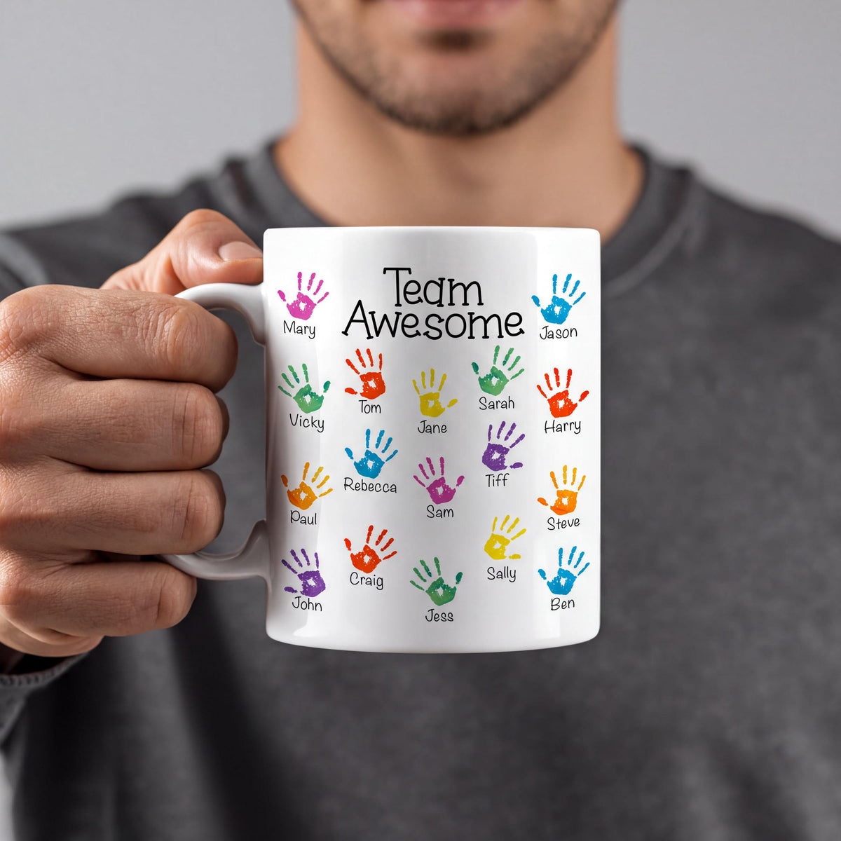 A person is holding a white mug with colorful handprints on it, each representing a different person's name.