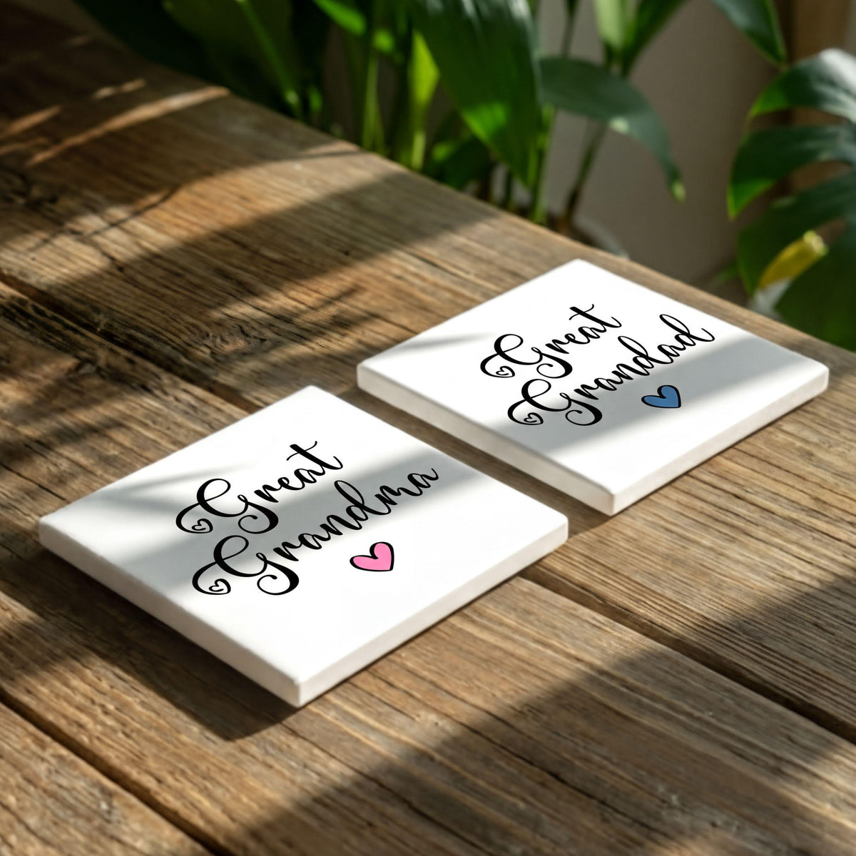 Two white coasters with the text "Great Grandma" printed on them, placed on a wooden surface with some greenery in the background.
