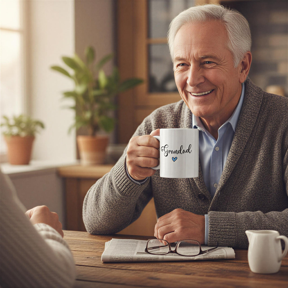 An older man with gray hair is sitting at a table, holding a mug with the word "Grandad" written on it. He is smiling and appears to be enjoying a moment of relaxation.