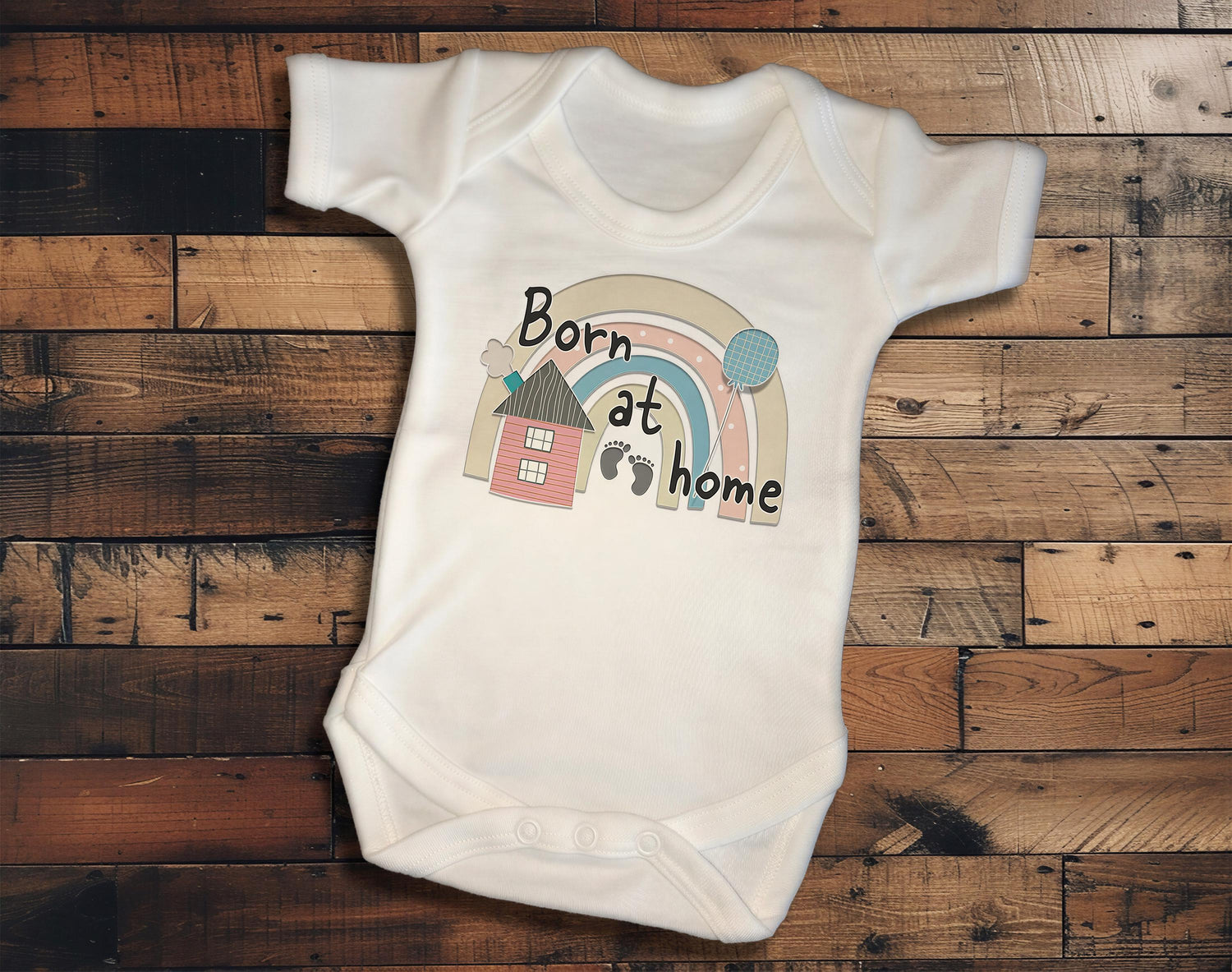 A white baby onesie with a rainbow graphic and the text "Born at home" printed on it, laid out on a wooden surface.