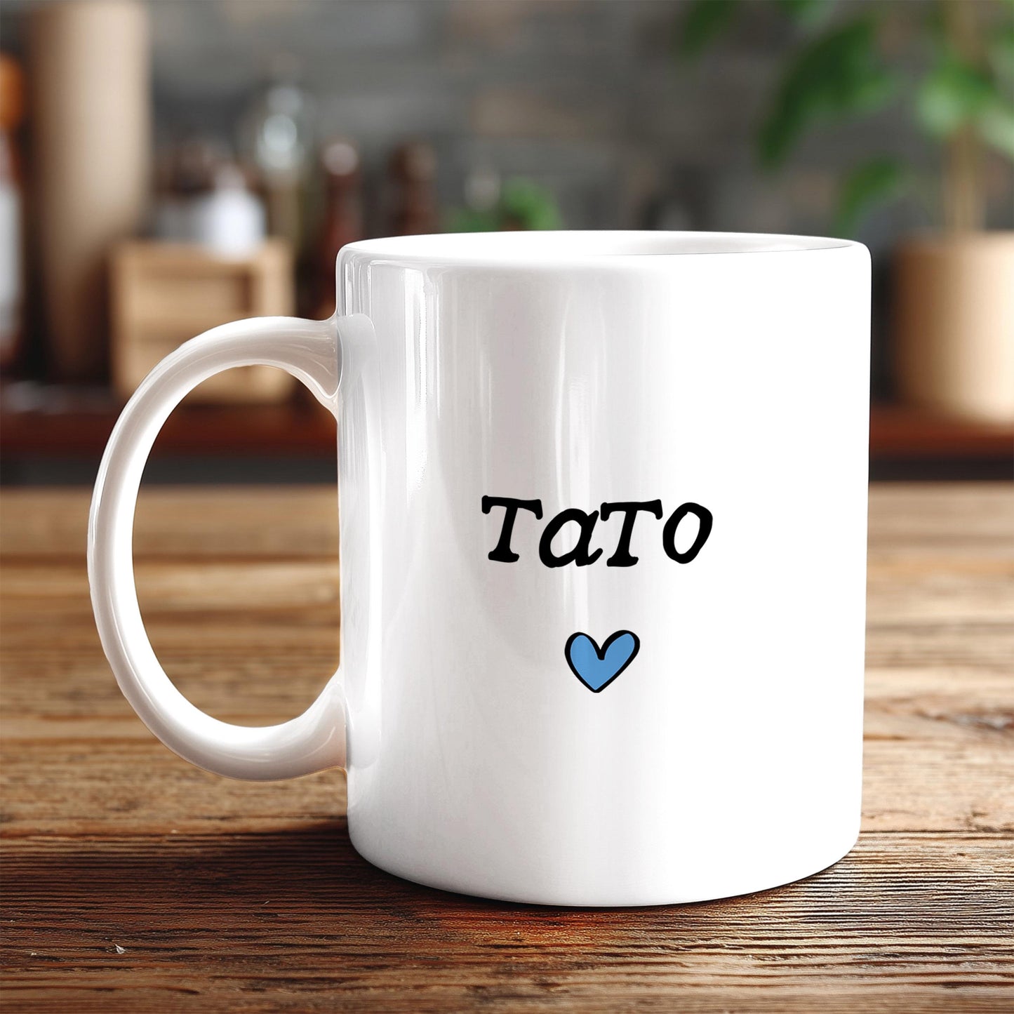 A white ceramic mug with the word "Tato" written on it, along with a small blue heart symbol, sits on a wooden surface.