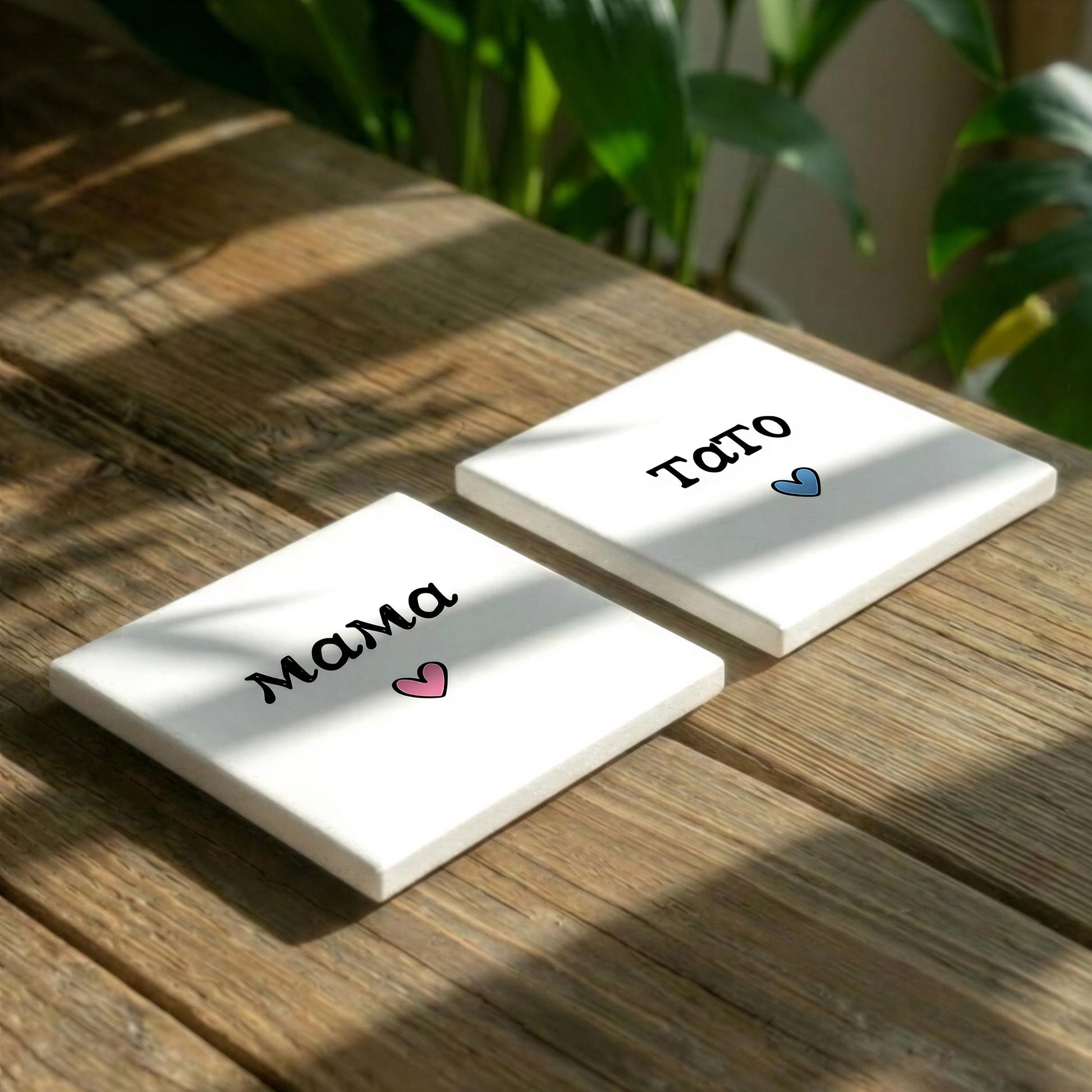 Two white square coasters with the word "Mama" and a heart symbol printed on them, placed on a wooden surface with plants in the background.