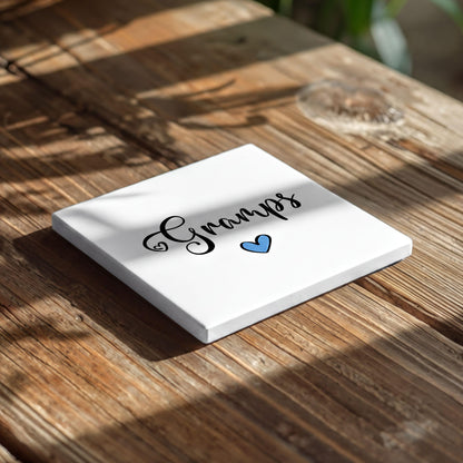 A white square coaster with the word "Grandpa" written on it, along with a blue heart symbol, placed on a wooden surface.