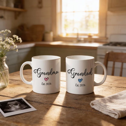 Two white mugs with the text "Grandma" and "Grandpa" printed on them, along with the year "Est. 2026" and a heart symbol, are placed on a wooden table. There is also a vase with flowers and a photograph of an ultrasound image in the background.
