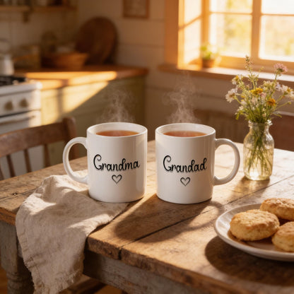 Two white mugs with the text "Grandma" and "Grandad" printed on them, along with a plate of cookies and a vase of flowers, are placed on a wooden table in a cozy kitchen setting.