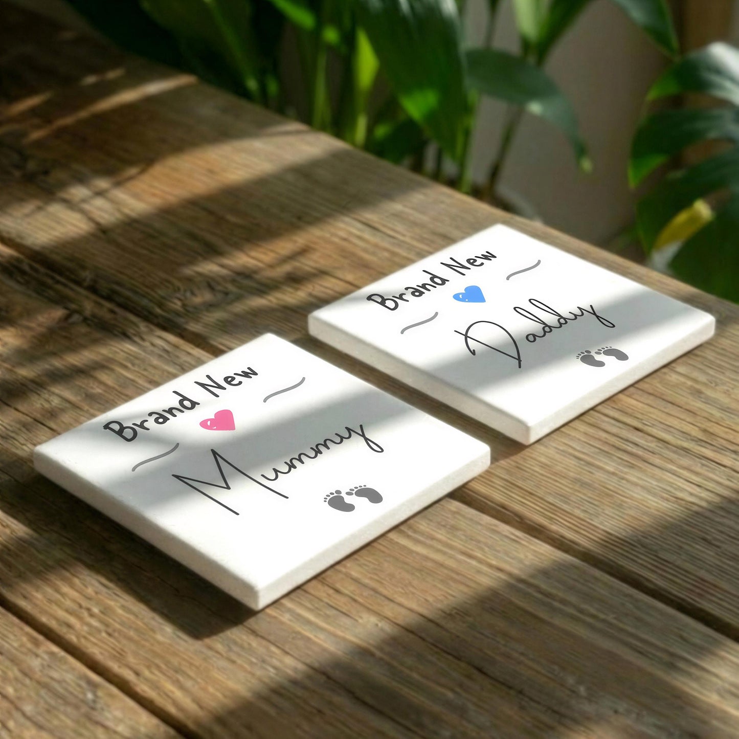 Two white coasters with text and illustrations on them, placed on a wooden surface with plants in the background.