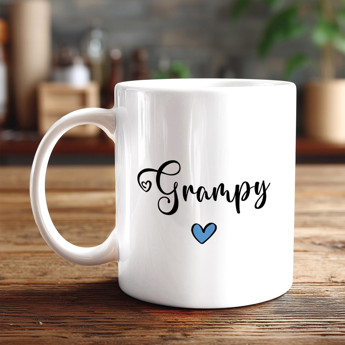 A white ceramic mug with the word "Grampy" written on it, along with a blue heart symbol, sits on a wooden surface.