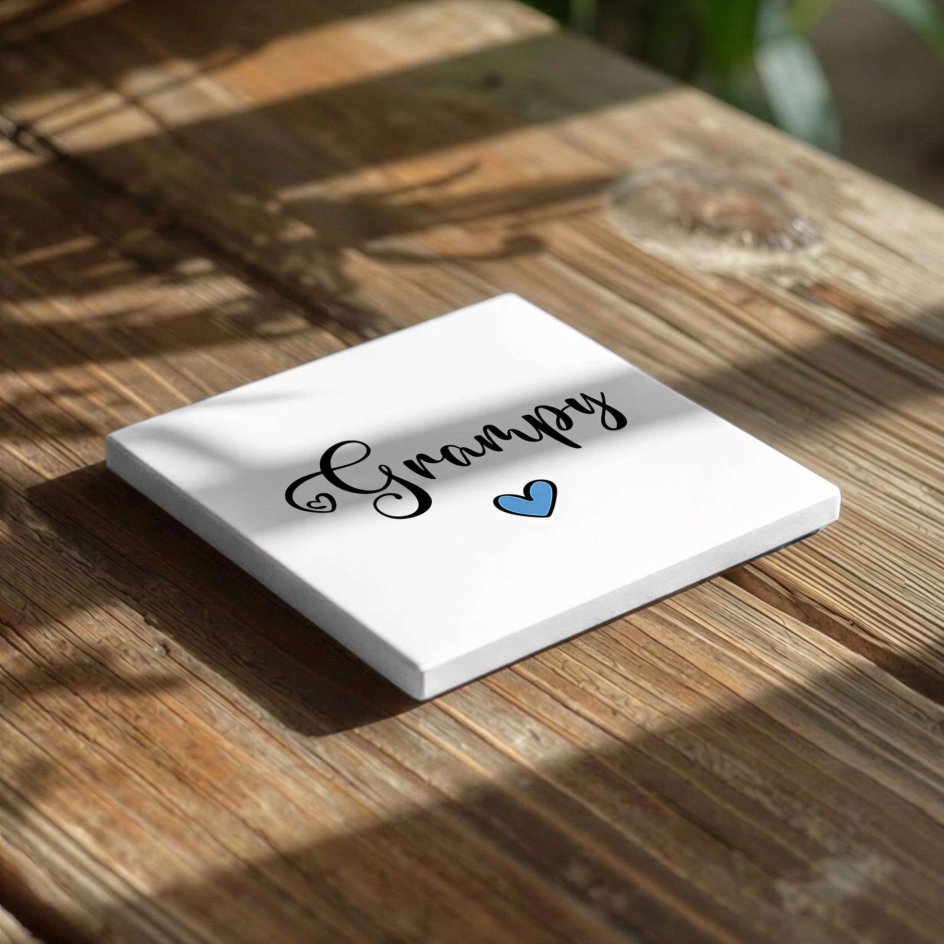 A white square coaster with the word "Grumpy" written on it, along with a blue heart symbol, placed on a wooden surface.