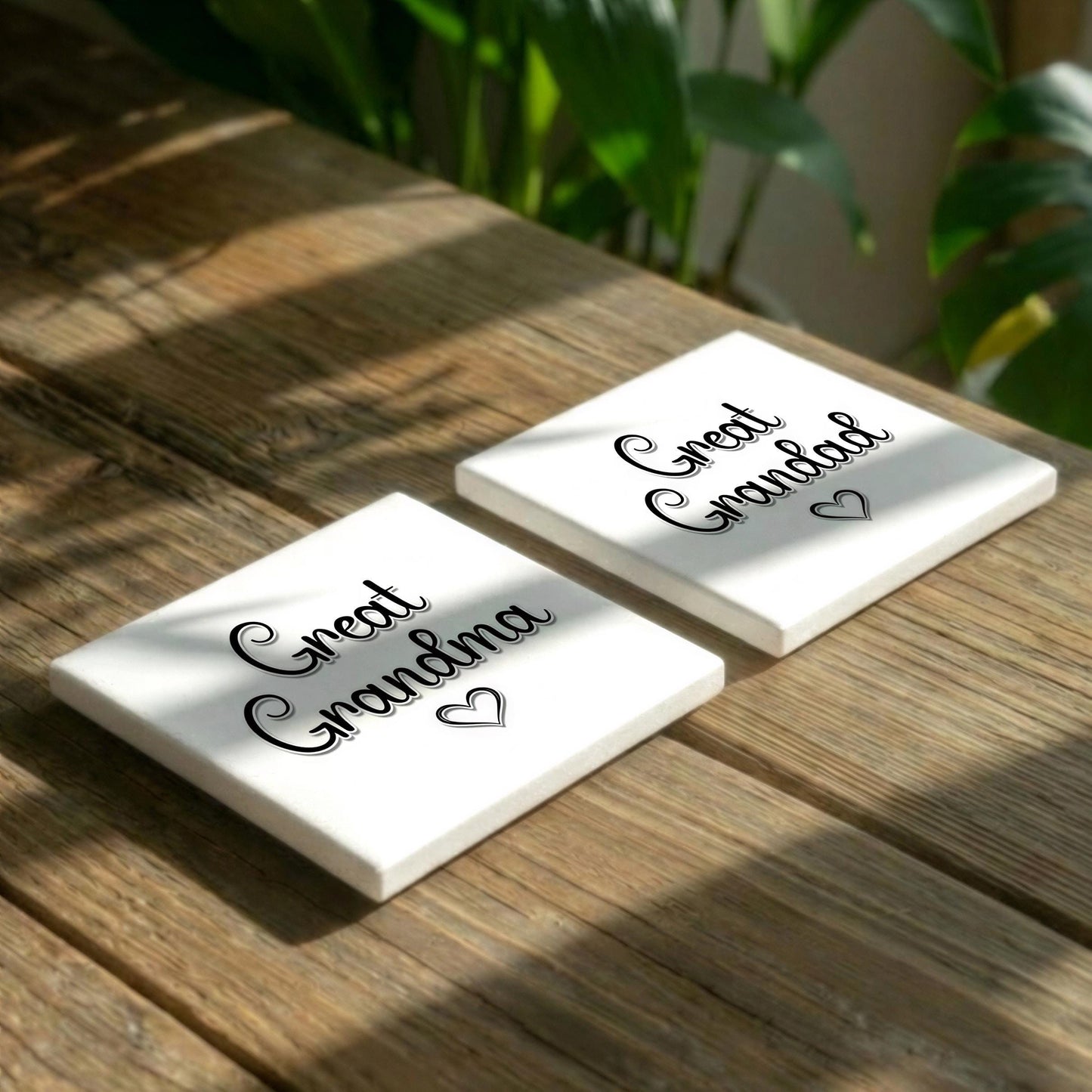 Two white square coasters with the text "Great Grandma" printed on them, placed on a wooden surface with some greenery in the background.
