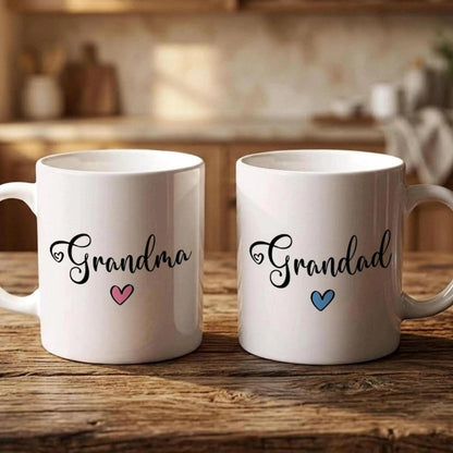 Two white mugs with the text "Grandma" and "Grandpa" printed on them, along with a heart symbol, are placed on a wooden surface.