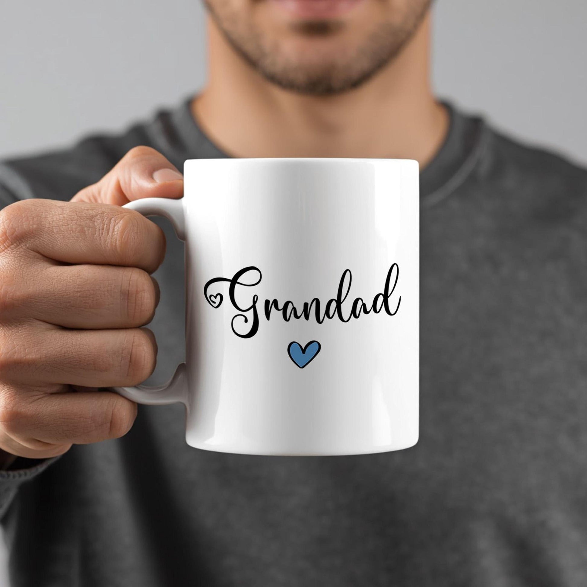 A person is holding a white mug with the word "Grandad" written on it in a cursive font.