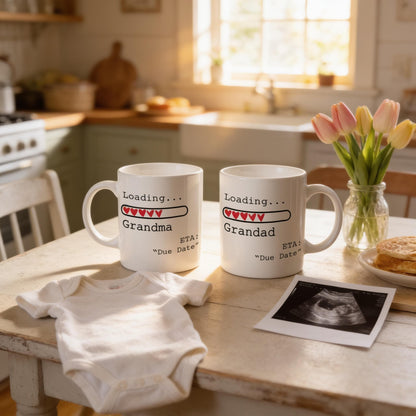 two white mugs with text that reads "Loading... Grandma" and "ETA: 'Due Date'" printed on them, placed on a kitchen counter next to a baby onesie, a vase of tulips, and an ultrasound image.