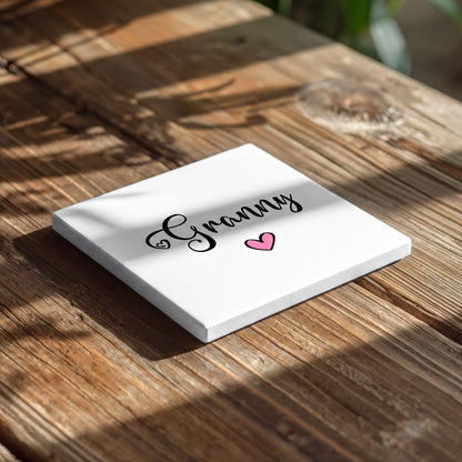 A white square coaster with the word "Granny" written on it, along with a pink heart symbol, placed on a wooden surface.