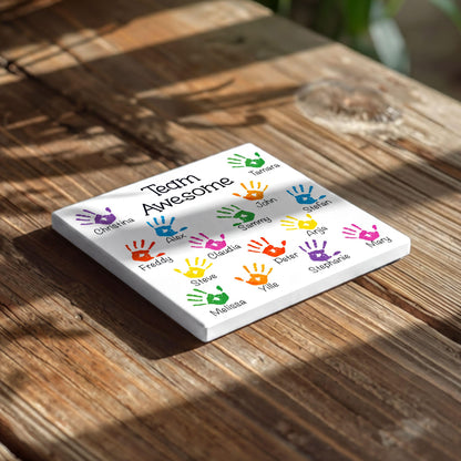 A white square coaster with colorful handprint designs and the text "Team Awesome" printed on it, placed on a wooden surface.