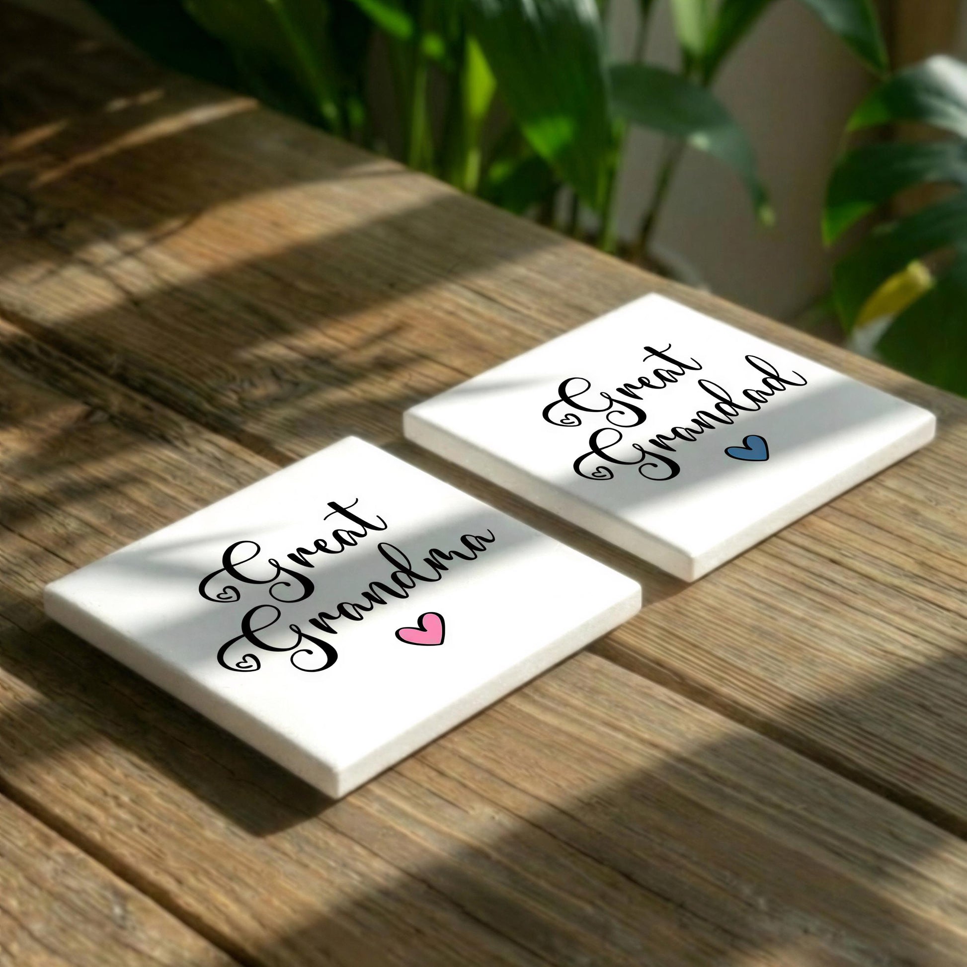 Two white coasters with the text "Great Grandma" printed on them, placed on a wooden surface with some greenery in the background.