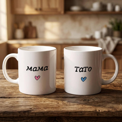 Two white mugs with the names "Mama" and "Tato" printed on them, along with a heart symbol, are placed on a wooden surface.