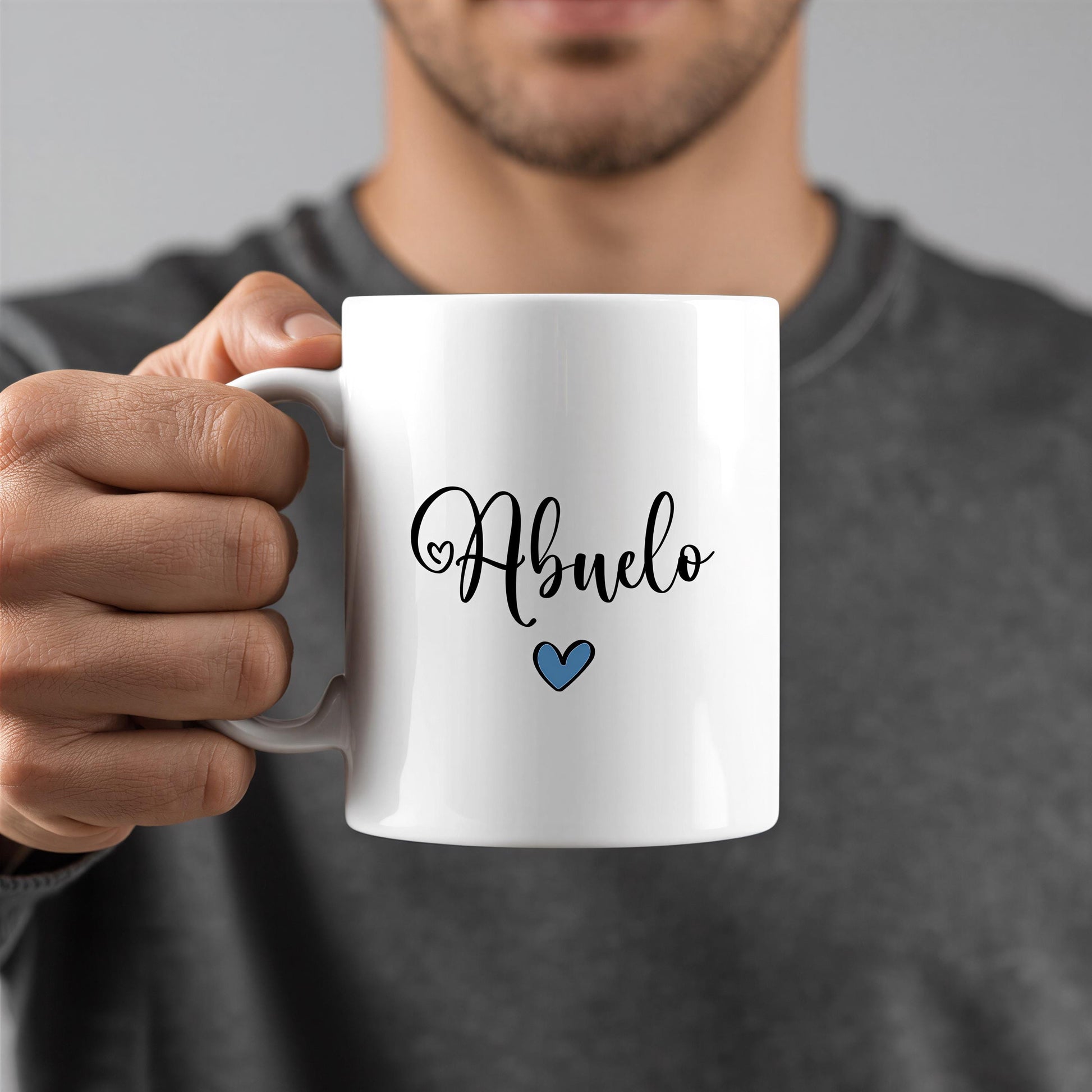 A person is holding a white mug with the word "Abuelo" written on it in a cursive font, along with a small blue heart symbol.