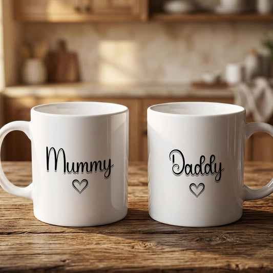 Two white mugs with black text that says "Mummy" and "Dadmy" on them, placed on a wooden surface.