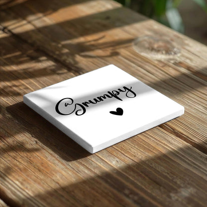 A white square coaster with the word "Grumpy" written on it, along with a small heart symbol, placed on a wooden surface.