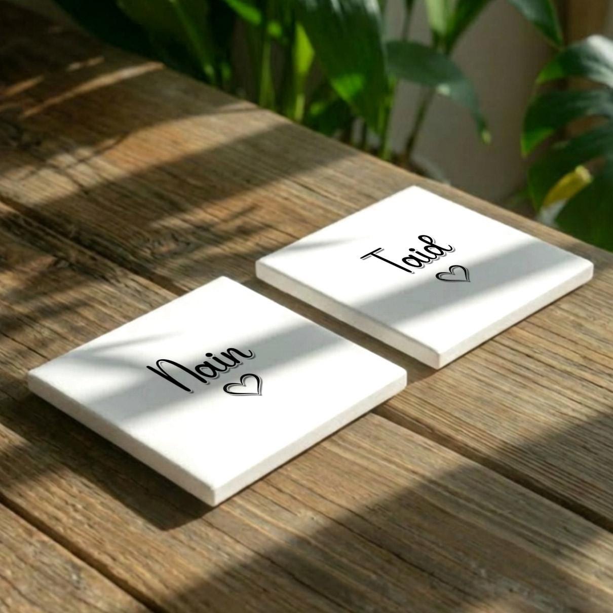 Two white square coasters with the names "Noir" and "Tail" written on them, along with a heart symbol, are placed on a wooden surface.