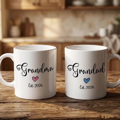 Two white ceramic mugs with the text "Grandma" and "Grandpa" printed on them, along with a heart symbol and the year "Est. 2026" below. The mugs are placed on a wooden surface, possibly a table or countertop.