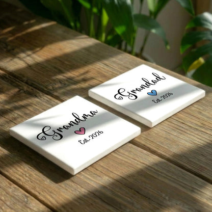 Two white coasters with the text "Grandma" and "Grandpa" printed on them, along with a heart symbol, are placed on a wooden surface.