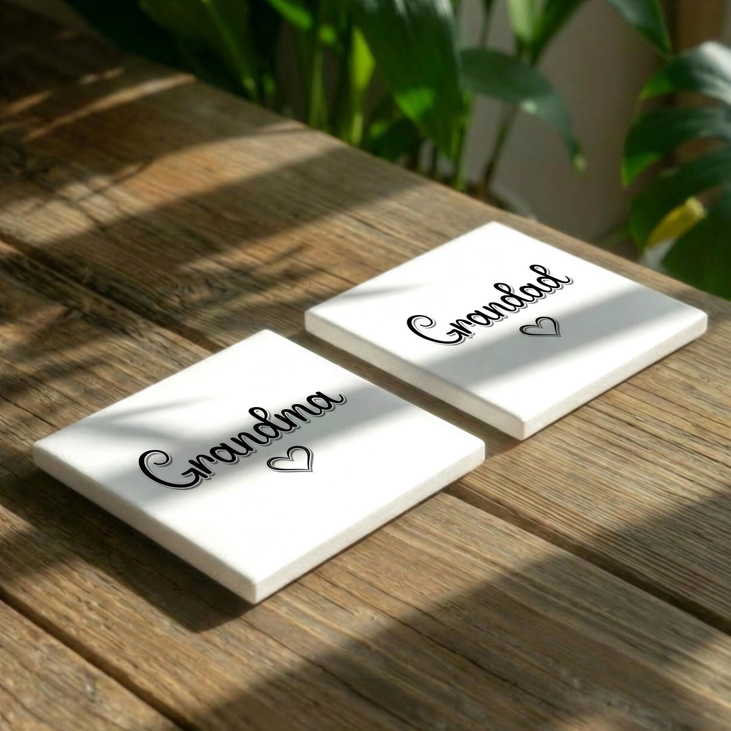 Two white square coasters with the word "Grandma" printed on them, placed on a wooden surface with green plants in the background.