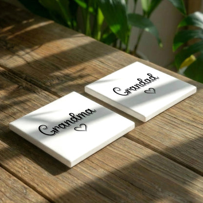 Two white square coasters with the word "Grandma" printed on them, placed on a wooden surface with green plants in the background.